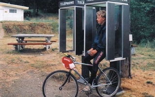 A boy on a bike leans against a telephone booth.