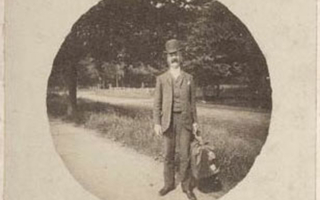 A man stands on the road holding a bag. Trees and grass are in the background.